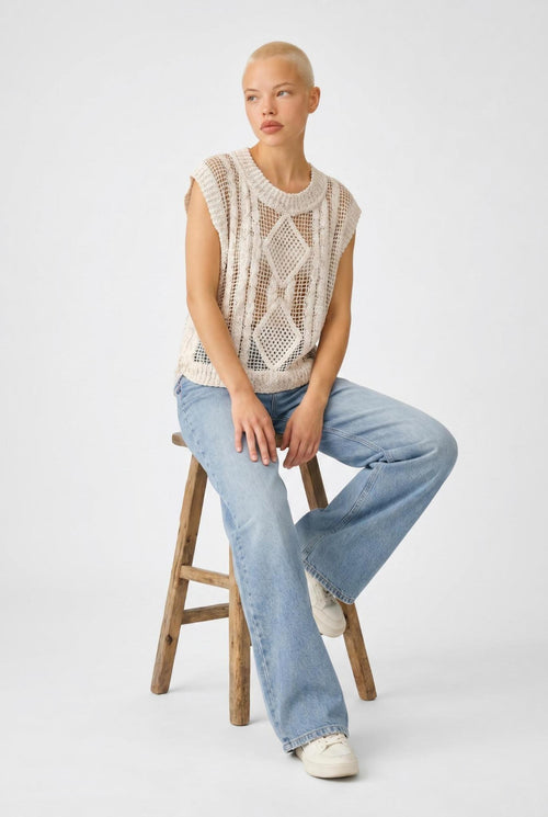 Woman sitting on a wooden stool wearing a sleeveless crochet top and jeans against a white background